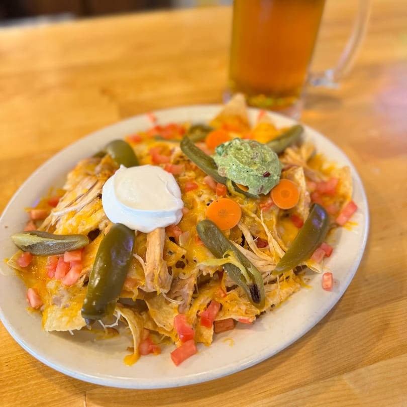 A plate of chicken nachos with cheese, tomatoes, jalapeños, sour cream, and guacamole, with a beer.