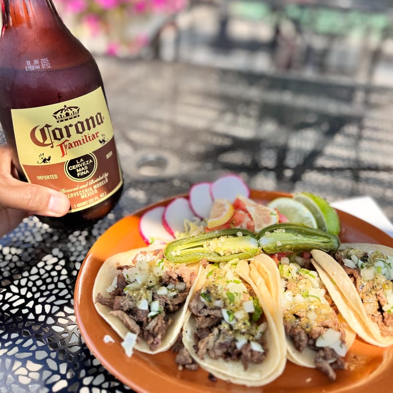 A hand holds a Corona Familiar beer bottle next to a plate of carne asada tacos with jalapeños.