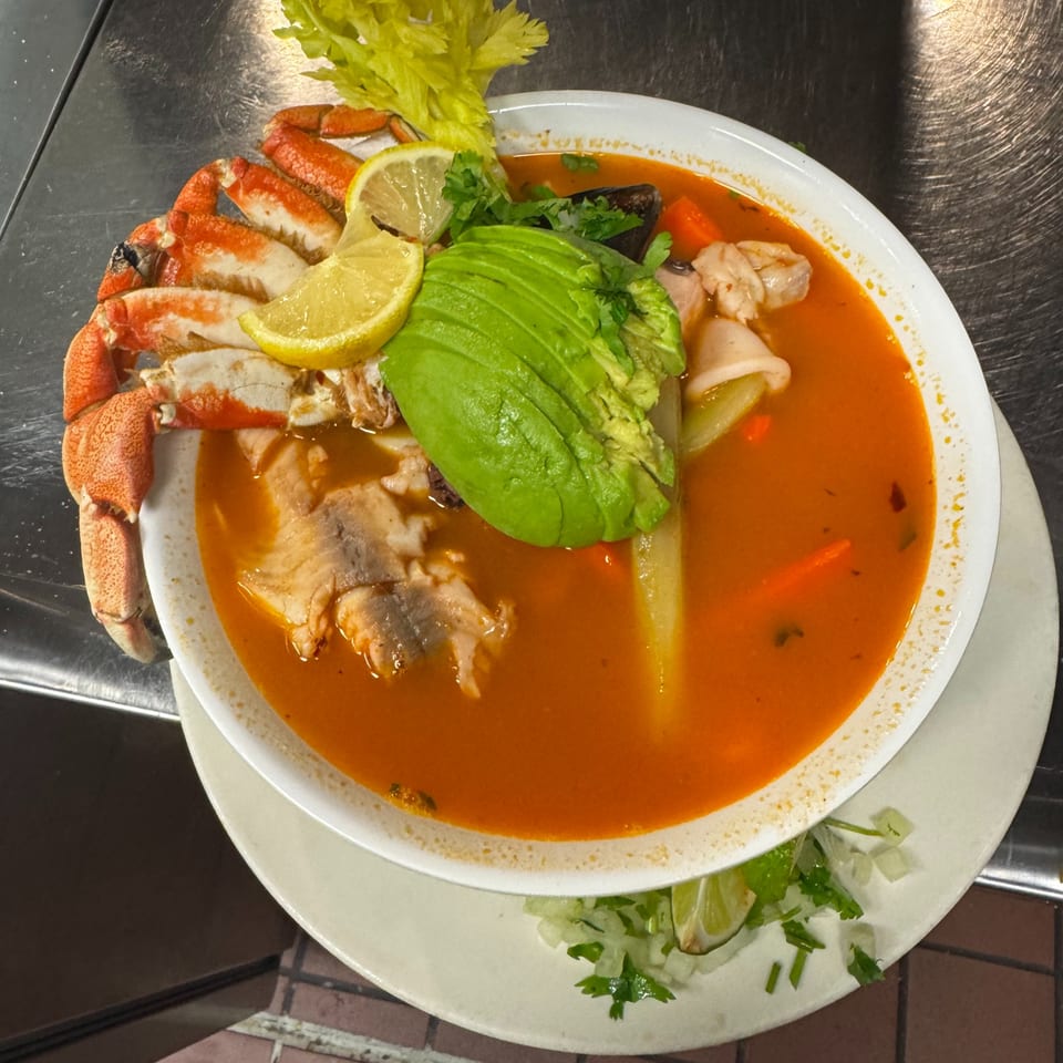 Mexican seafood soup with crab legs, fish, avocado, and lemon slices in a white bowl.