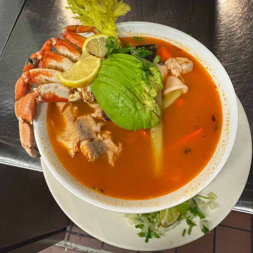 Mexican seafood soup with crab legs, fish, avocado, and lemon slices in a white bowl.