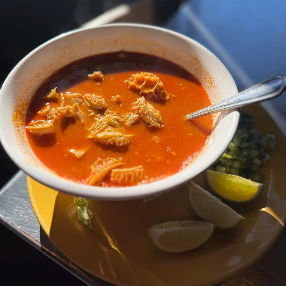Menudo soup in a white bowl with lime wedges and chopped onions on a yellow plate.