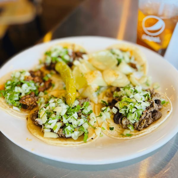 A plate of five Mexican street tacos with meat, cilantro, and onions, alongside a pickled pepper.