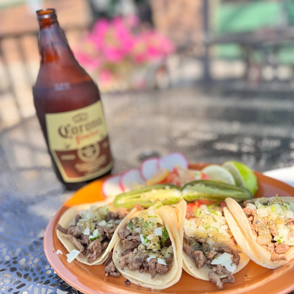 Three street tacos with meat, onions, and cilantro, served with a Corona beer bottle.