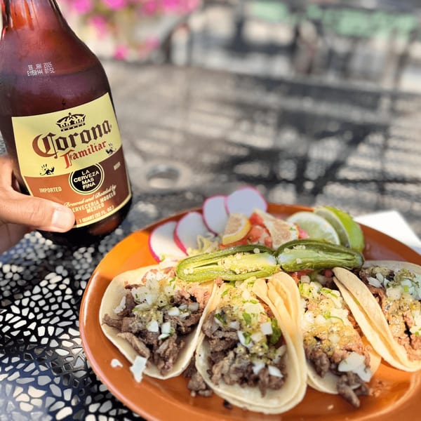 A hand holding a Corona Familiar beer bottle next to a plate of carne asada tacos with toppings.