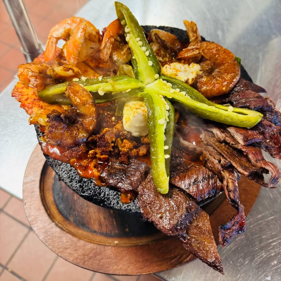 Sizzling molcajete with shrimp, steak, chorizo, cheese, and jalapeños, served on a wooden base.