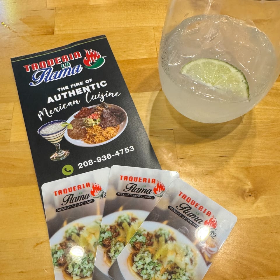 Taqueria La Flama menu, business cards, and a drink with a lime wedge on a wooden table.