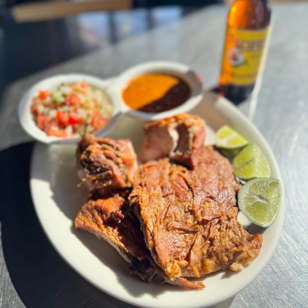 Crispy carnitas with lime wedges, pico de gallo, and two sauces on a white plate with a beer.