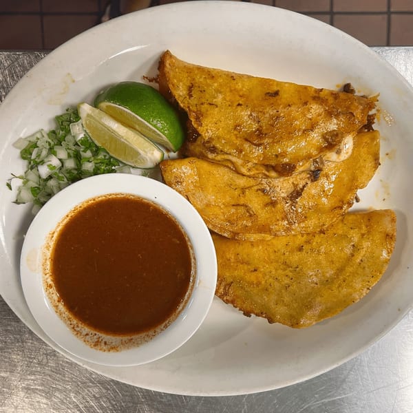 Three golden-brown birria tacos with lime wedges, chopped onions, cilantro, and a side of consommé.