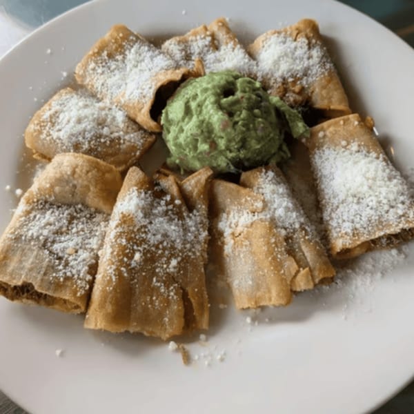 Crispy flautas dusted with cotija cheese, surrounding a scoop of guacamole on a white plate.