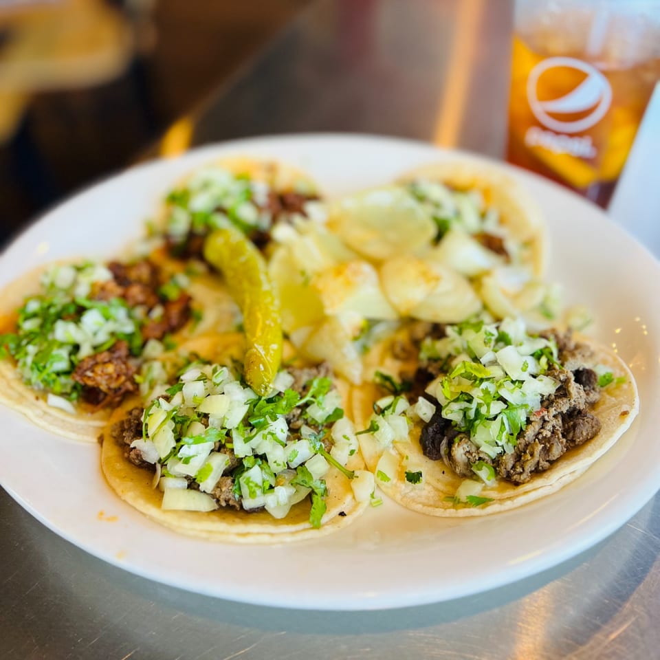 Five Taqueria La Flama tacos with meat, cilantro, and onions, plus a pepper and drink.